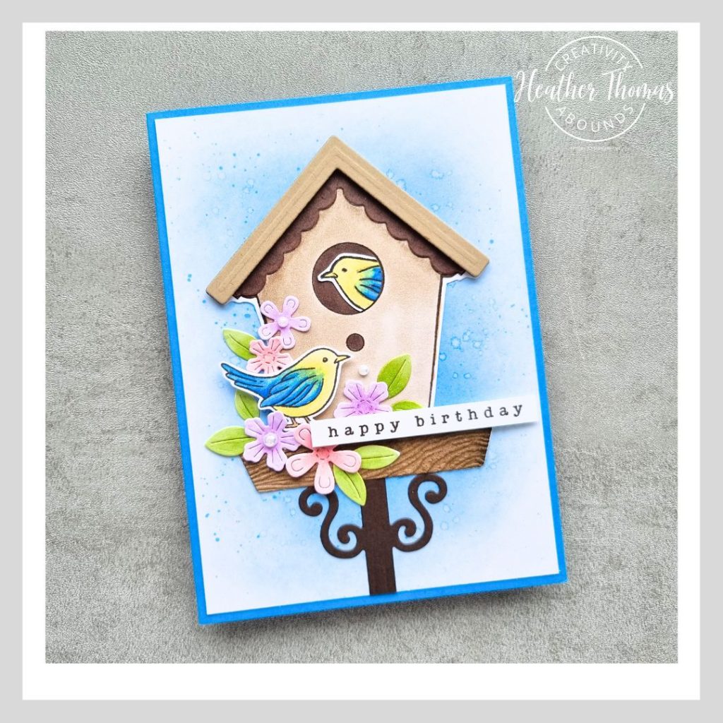 A handmade card with a bird house on an ink blended background, with two birds, some flowers and the sentiment Happy Birthday.