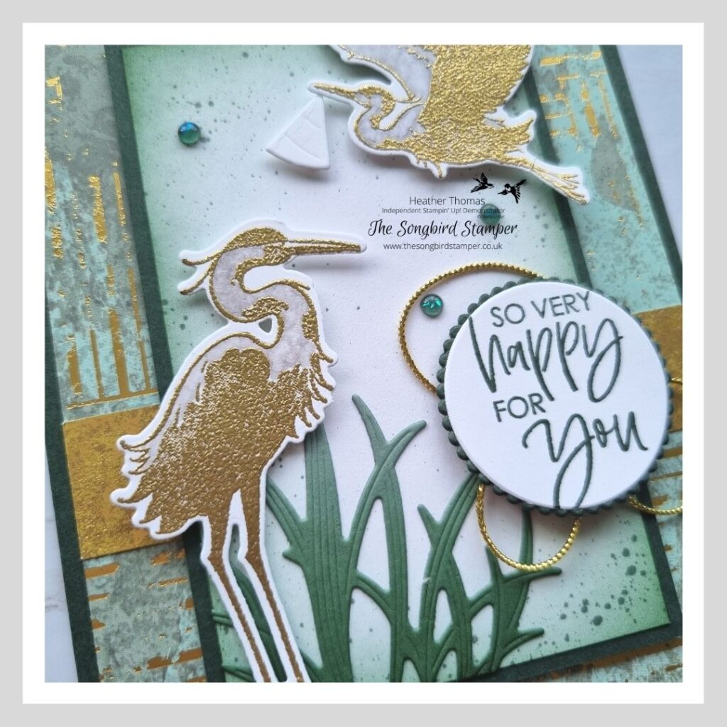 A close up of A new baby card with a twist, in green and gold with herons carrying a baby sling